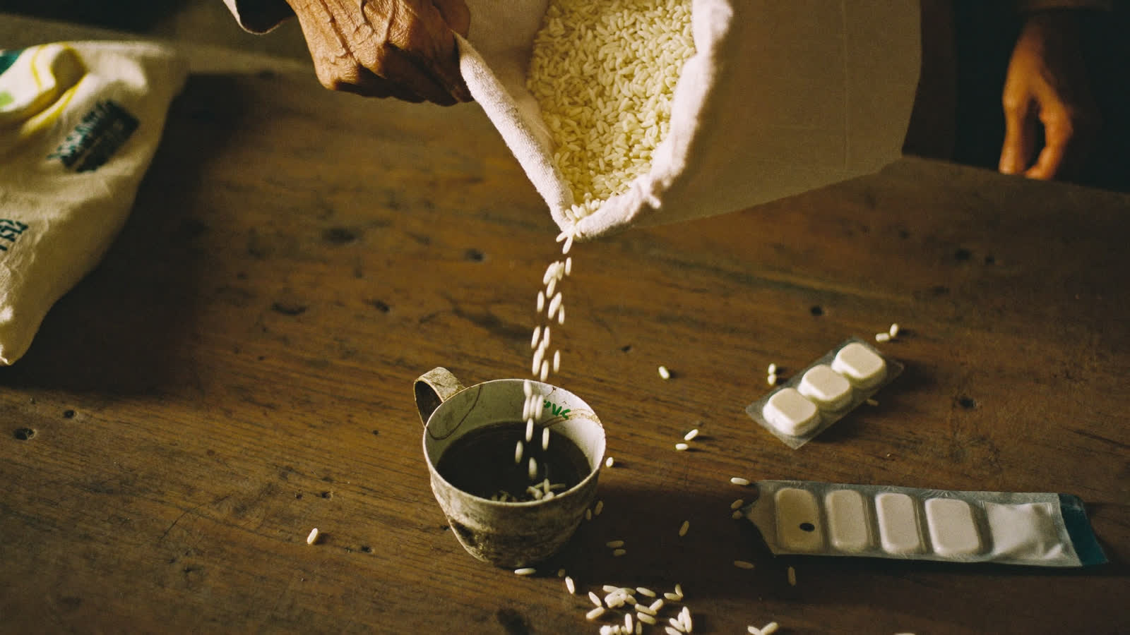Rice pouring from a cloth sack into a tin measure on a wooden table, beside a strip of tablets.