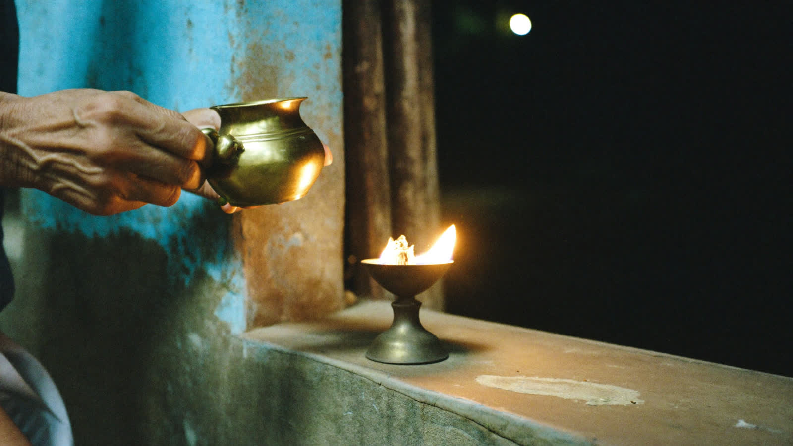Hands tipping oil toward a small brass lamp whose flame has just caught, the only light in the dark.